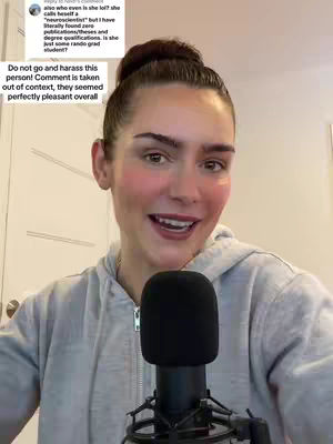 A woman with brown hair pulled up in a bun is centered in the frame, smiling and holding a black microphone. She wears a light gray hoodie. Text including a comment and the phrase 'Do not go and harass this person!' is overlaid near the top left.