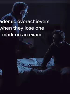 A dark, indoor scene featuring two silhouetted figures. One figure, in the foreground, appears to be standing and gesturing, while the other sits on a bed, looking down. The lighting is dim, with a cool blue tone, suggesting a night setting. The text overlay indicates a theme of academic stress.