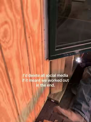 The image shows a close-up of a wooden wall with a window frame and a figure partially visible in the corner. The wall is made of vertical wooden planks, displaying a natural wood grain and a warm, orange hue. A dark green window frame cuts across the upper right corner of the image. The text overlayed reads, "I'd delete all social media if it meant we worked out in the end."