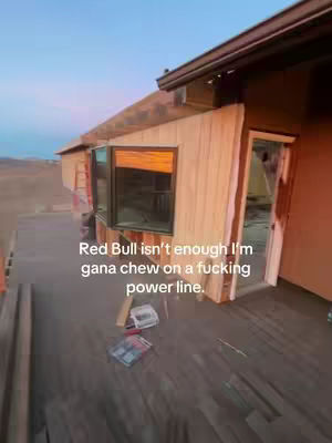 The image shows an exterior view of a building under construction. The building is made of wood and has a dark roof. A window is visible, and the sky is a gradient of blue and pink. The image is taken from a low angle, looking up at the building.