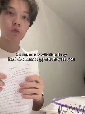 A young man in a white t-shirt is holding a paper with handwritten notes, looking pensively at the camera. Text overlay says, 'Someone is wishing they had the same opportunity as you'. The background suggests an indoor setting, lit by natural light, with a desk and notebooks partly visible.