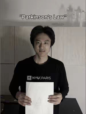 A man stands in front of what seems like a white wall with a paper at the bottom half that has a triangle drawing and the words "Parkinson's Law" in a white text overlay above him, in a room.