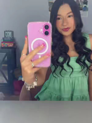 A selfie-style image featuring a young woman holding a pink phone case. She has long, dark, wavy hair and is wearing a green top. Her nails are painted pink. The background is a blurred room.