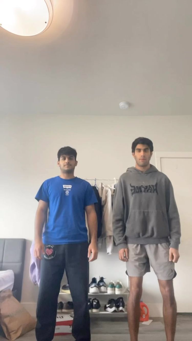 Two young men are standing in what appears to be a living space. The man on the left is wearing a blue t-shirt and black sweatpants, and the one on the right is wearing a gray hoodie and gray shorts. They are both standing in front of a plain white wall. In the background, there is a closet with some clothes on hangers and a shelf with shoes.