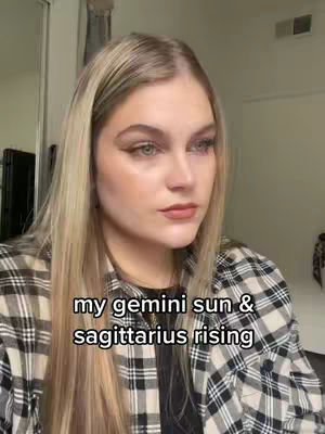 A medium shot shows a young woman with long blonde hair and a plaid overshirt. She is looking slightly to the right of the camera with a contemplative expression. Overlayed text reads 'my gemini sun & sagittarius rising'. The background is blurred, suggesting an indoor setting.
