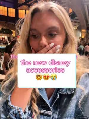 A selfie-style shot featuring a young woman with blonde hair, covering her mouth with her hand, wearing a denim jacket. A rectangular text overlay near her face reads 'the new disney accessories', with emoji reactions below. The background is softly blurred, showing an interior setting, possibly a store or event space.