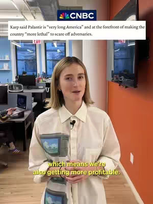 A woman in a white button-down shirt stands in an office setting, with a CNBC logo and text overlayed. She smiles and speaks to the camera, with a background featuring computer screens and windows.
