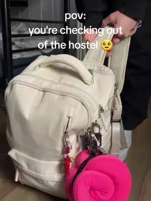 A person holding a white backpack with a white strap, a pink item hanging from it, against a background that appears to be a hallway or a room with dark objects.. The backpack has a few charms hanging from it. Text overlayed reading 'pov: you're checking out of the hostel' and a smiley face.