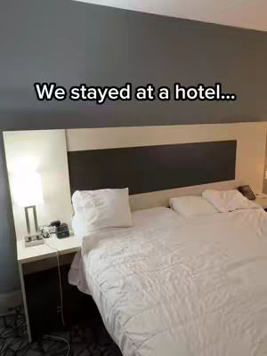 A hotel room with a bed and a lamp on a nightstand. The bed has white sheets, and there's a headboard on the wall.