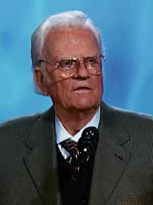 The image features a medium shot of an elderly man with white hair and glasses, likely in a studio setting. He is wearing a suit jacket and appears to be speaking. A microphone is positioned in front of him, suggesting he's delivering a message or presentation. The background is a plain blue color, which provides a clean backdrop, highlighting the man. The lighting seems to be even, illuminating his face.