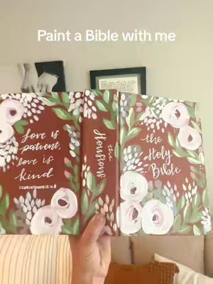 A person holds open a Bible that has been hand-painted with floral and script elements. The background includes a partial view of a light-colored wall with framed artwork and a window. The Bible cover displays the words 'Love is patient, love is kind' on one side and 'the Holy Bible' on the other. The color palette is warm, utilizing shades of brown, green, and pink with white.
