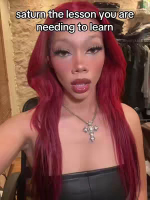 A woman with long red hair and full makeup is centered in a video selfie. The background is neutral, with a glimpse of clothing racks and a light-colored wall. She wears a black strapless top and a cross pendant while looking at the camera.