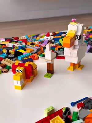 An overhead shot of several Lego animal figures sitting on a white surface. To the left, a yellow lion is depicted, and in the center are two bird-like figures; one is a goose, and the other is a larger white bird. A wide array of colorful Lego bricks are spread out in the background, suggesting a construction zone. The scene focuses on play and creativity with the Lego blocks.