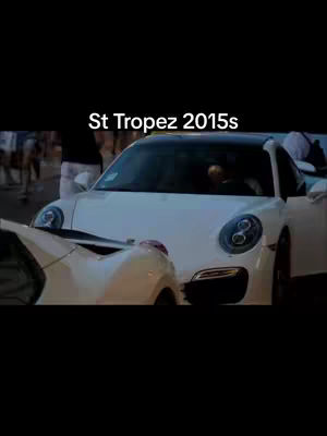 The image shows two white sports cars parked closely together, with their front ends visible. The car in the foreground has its headlights facing the viewer, and appears to be a Porsche. The background is blurry, but indicates a place with people likely St. Tropez
