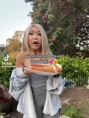 A woman with blonde hair is holding a plate with a hotdog on it. She is standing in front of foliage. Text over the image says 'jennifer and david bonding!'
