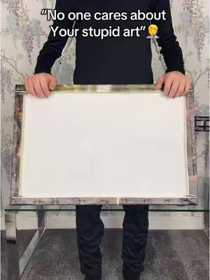 A person holding a white canvas in front of them, with text overlaid at the top of the image. The person is dressed in a black long-sleeved shirt and black pants. The setting appears to be indoors, with a patterned wallpaper visible in the background.