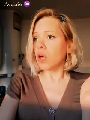 A blonde woman with a slightly surprised expression on her face, seen from the chest up. The background is blurred, with hints of a room and some light. The woman has light skin, short blonde hair, and is wearing a dark brown top.