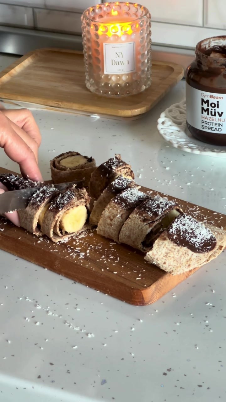 A close-up shot of a wooden cutting board with several sliced wraps filled with a light-colored cream and banana slices. Dark brown spots of a topping are scattered over the wraps with white powder and a jar of hazelnut protein spread is in the background. The background features a lit candle.