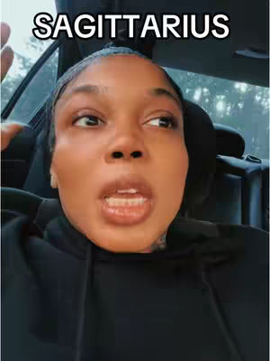 A selfie-style shot featuring a woman with dark skin in a car interior, focusing on her face and upper body. Above her head, the word "SAGITTARIUS" is displayed in all caps within a white rounded rectangle. She appears to be speaking, with her mouth open. The car's interior is dark, with seat details visible behind her. Daylight comes from the windows, casting shadows and reflections on her face.