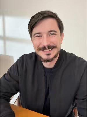 The cover image features a close-up of a smiling man. He has short, dark hair, a mustache, and a neatly trimmed beard. He's wearing a black bomber jacket over a black t-shirt. The background is a softly lit, solid, light color.
