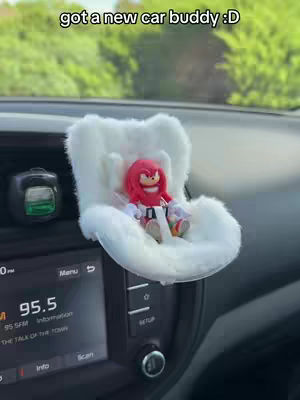 The interior of a car is shown, focusing on the dashboard and the radio display. A novelty seat, shaped like a fluffy white cloud, is installed. Sitting in the seat is a Knuckles the Echidna figurine. The text overlay states, "got a new car buddy :D". The background includes a view of green foliage outside the car window.