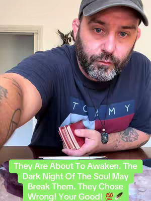 A selfie-style video cover image with a man holding a stack of tarot cards. The man is wearing a dark baseball cap, a navy blue t-shirt with white and red stripes. A black necklace with a green stone hangs around his neck. The background is a light wall with a window. Text overlayed in a light green box on the bottom of the image provides the content's hook.