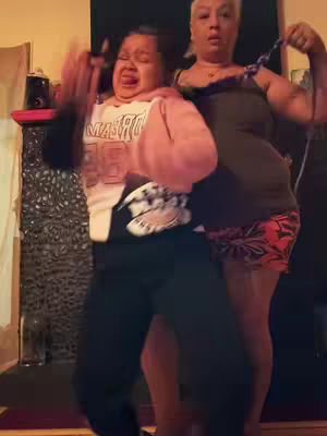 Two women dancing in a room with a dark background.  One appears to be holding the other in a neck-hold while they make exaggerated facial expressions.