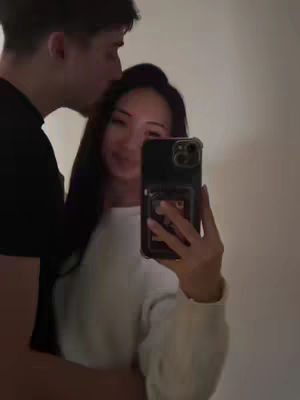 A selfie taken in front of a mirror, showing a young woman in a white sweater embraced by a man. The woman is holding the phone, capturing their faces and the surrounding environment, while the man kisses her on the head. The composition is intimate with a neutral and illuminated background.