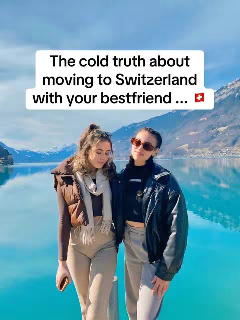 Two young women stand side-by-side in front of a picturesque lake and mountain backdrop. They are smiling, with one wearing sunglasses and a black jacket over a black shirt and gray pants, and the other wearing a brown vest with a scarf, long-sleeved top, and light-colored pants. The framing is from the waist up.