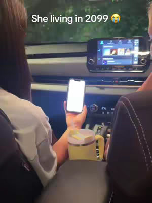 A person is sitting in a car, holding a phone with a blank white screen. A yellow mug is in the center console. Outside, the car environment is partially obscured by the darkness. The overlay text reads: She living in 2099 with a shocked face emoji.