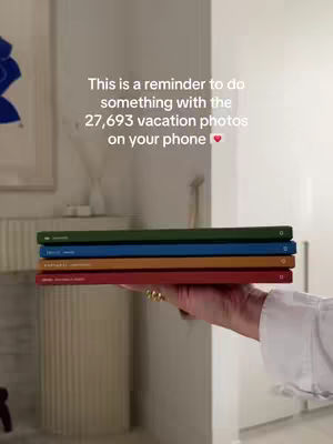 A hand holding a stack of colorful photo books, on top of those books is a text overlay, set against a blurred background of a light-colored wall and some decor.