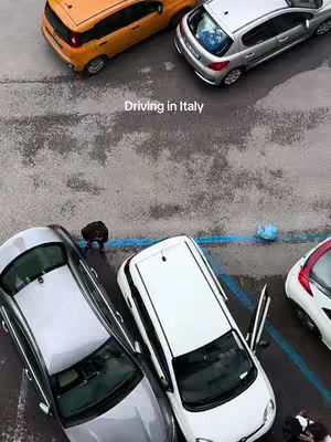 An overhead shot of parked cars in a lot with text overlayed. The cars are a mix of silver, orange and white parked on an asphalt surface with blue lines. The sky is overcast.