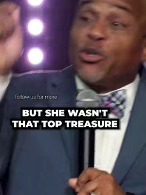A medium shot features a Black man speaking with a microphone in his right hand. He's wearing a dark suit with a white collared shirt and a patterned bow-tie. The background shows a blurred purple and white lights. Text overlays in bold, white letters reading "BUT SHE WASN'T THAT TOP TREASURE" appear over the image.