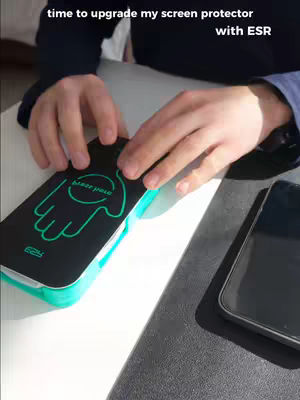The image shows a person's hands partially visible, centered on the frame, interacting with protective packaging on a white surface. Above the hands is some text, on the left side of the image, the screen protector packaging is visible, and next to it, is a black phone with a gray border.