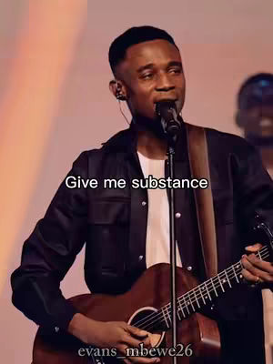 A man with short black hair is playing a brown acoustic guitar and singing into a microphone. The background is softly lit with warm tones. Text overlay across the image reads 'Give me substance.'