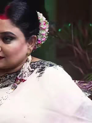 A close-up shot features a woman. Her dark hair is styled up with a bun with flowers in the back. She has dark eye makeup and red lipstick. She wears a white top with a patterned neckline, and her skin appears fair. The background is blurred.