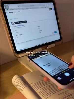 The image shows a setup for studying, with a tablet displaying a website and a phone positioned to scan a book page. A physical book is open on the desk. The overall composition suggests a study environment, utilizing both digital and physical resources.