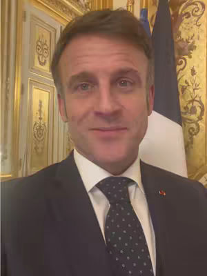 A selfie-style video featuring a man in a dark suit and tie, likely indoors. The man looks directly at the camera with a slight smile. Behind him, a French flag and ornate wall paneling suggest an official setting.