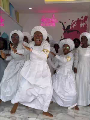 A group of people, dressed in white garments and hats, are standing and dancing in a brightly lit room. They are smiling and have their arms spread out. A neon sign in the background adds a pop of color.