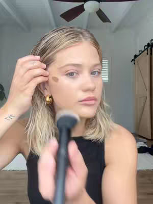 A woman with blonde hair is shown from the chest up, holding a makeup brush. She is applying foundation to her cheek, revealing a partially applied makeup look. The background has a ceiling fan and a door frame.