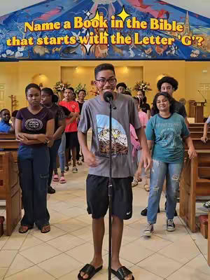 A young man stands in front of a group of people with a microphone in a church setting, a banner hanging above him with a trivia question. The man is wearing a gray t-shirt, black shorts, and black sandals. The background includes a group of people, wood pews, and decorative banners.