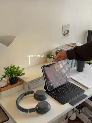 A well-lit desk with a laptop, headphones, and a small plant. The laptop displays a screen, and the headphones sit next to it. A keyboard and other items are also on the desk, creating a minimalist yet functional workspace. Text overlayed reads 'DAILY VLOG'.