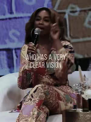 A woman with brown hair, wearing a patterned dress, is speaking into a microphone while seated. Text overlaid on the image says, "WHO HAS A VERY CLEAR VISION."