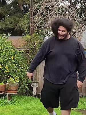 A man with dark hair and an afro walks in a backyard, smiling while looking downwards. He wears a black long-sleeve shirt and black shorts, the backyard includes green grass, foliage, and a fence in the background.