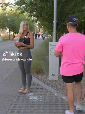 A woman in athletic wear stands facing the camera while holding something in her hands. Behind her is a person in a pink shirt with a baseball cap walking away. The background features a walkway lined with trees and other people.