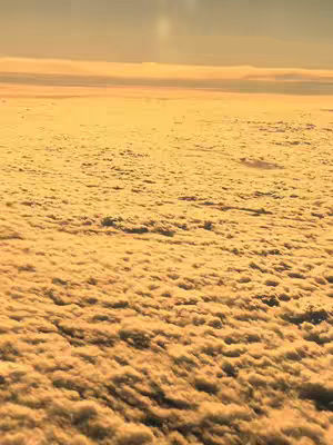 An aerial view of a dense cloud formation, with golden sunlight illuminating the scene. The clouds appear textured and layered, filling most of the frame. The sky above has a gradient of colors from light orange to a pale yellow.
