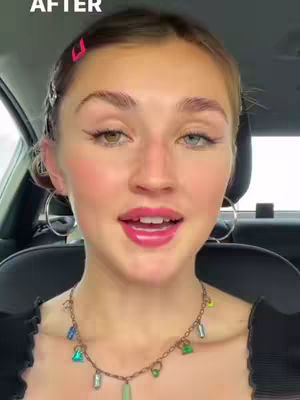 A close-up shot of a young woman in a car. She has a bright pink hair clip, and circular earrings. She has pink lipstick and her eyes are looking forward. She is wearing a jeweled necklace. The car interior is visible in the background.