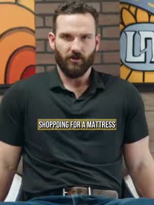 A medium shot of a man in a black polo shirt, seated and looking at the camera. Behind him is a split backdrop of a brick wall and abstract colorful shapes. Text overlayed reads 'SHOPPING FOR A MATTRESS'.