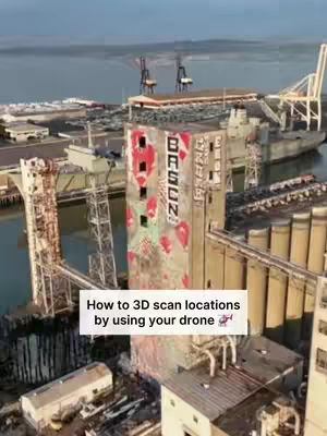 An aerial shot of an industrial waterfront area showcasing a towering, multi-story building with faded signage and several cylindrical storage tanks beside it. The scene captures a working harbor, with other structures, cranes, and the sea in the background, all bathed in daylight.
