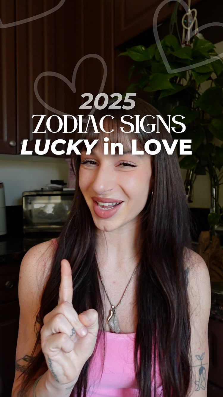 A woman with long dark hair and a pink top is smiling while pointing her index finger up. Overlaid on the image are white heart shapes and the text '2025 ZODIAC SIGNS LUCKY in LOVE'.